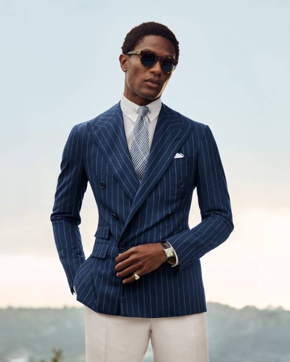 Black male model in old money style, double breasted navy blazer, repp tie, white oxford shirt, classic leather strap watch, silk picket square