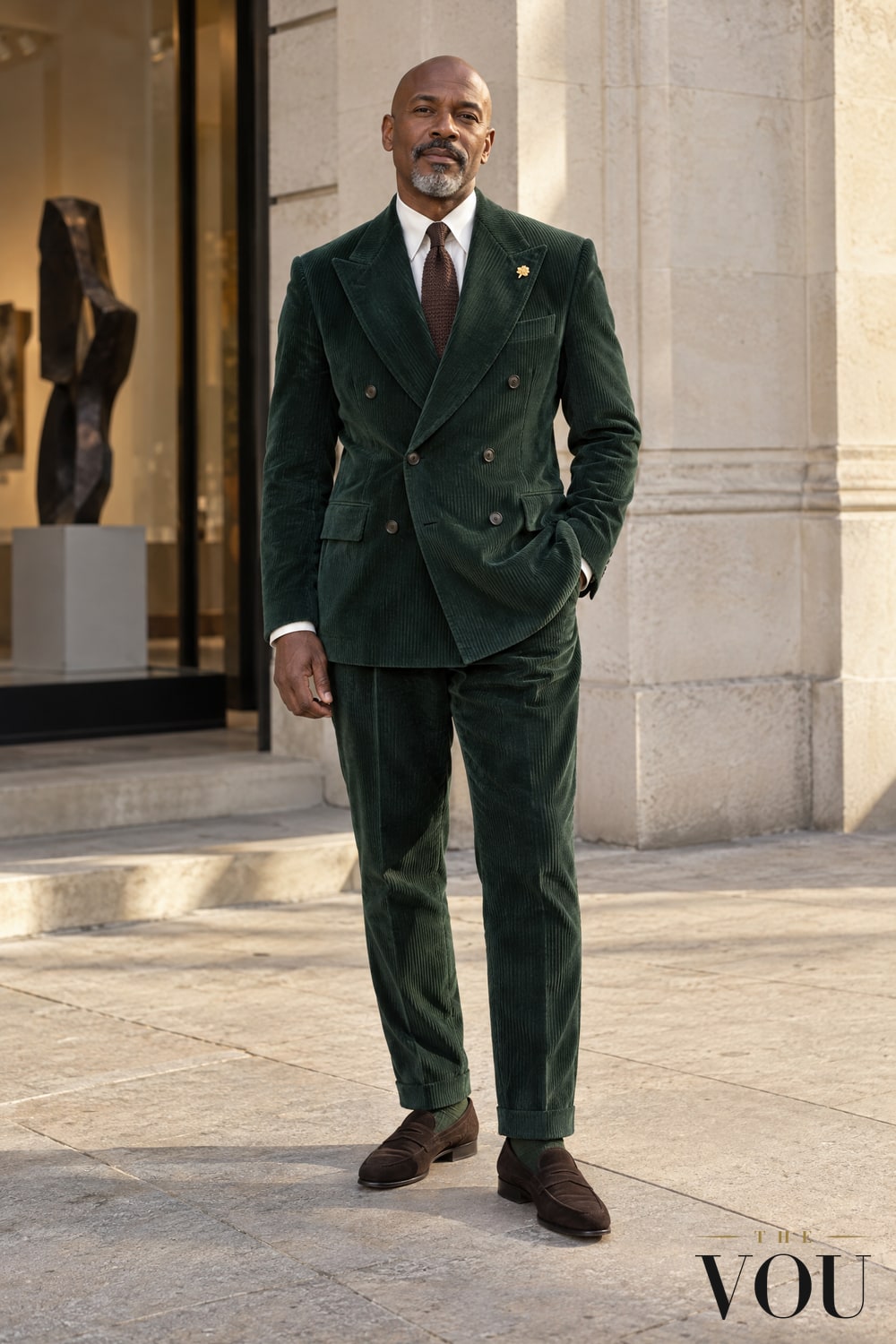 A Black man wearing a green double-breasted corduroy suit, ivory shirt, brown knitted tie and brown suede loafers.