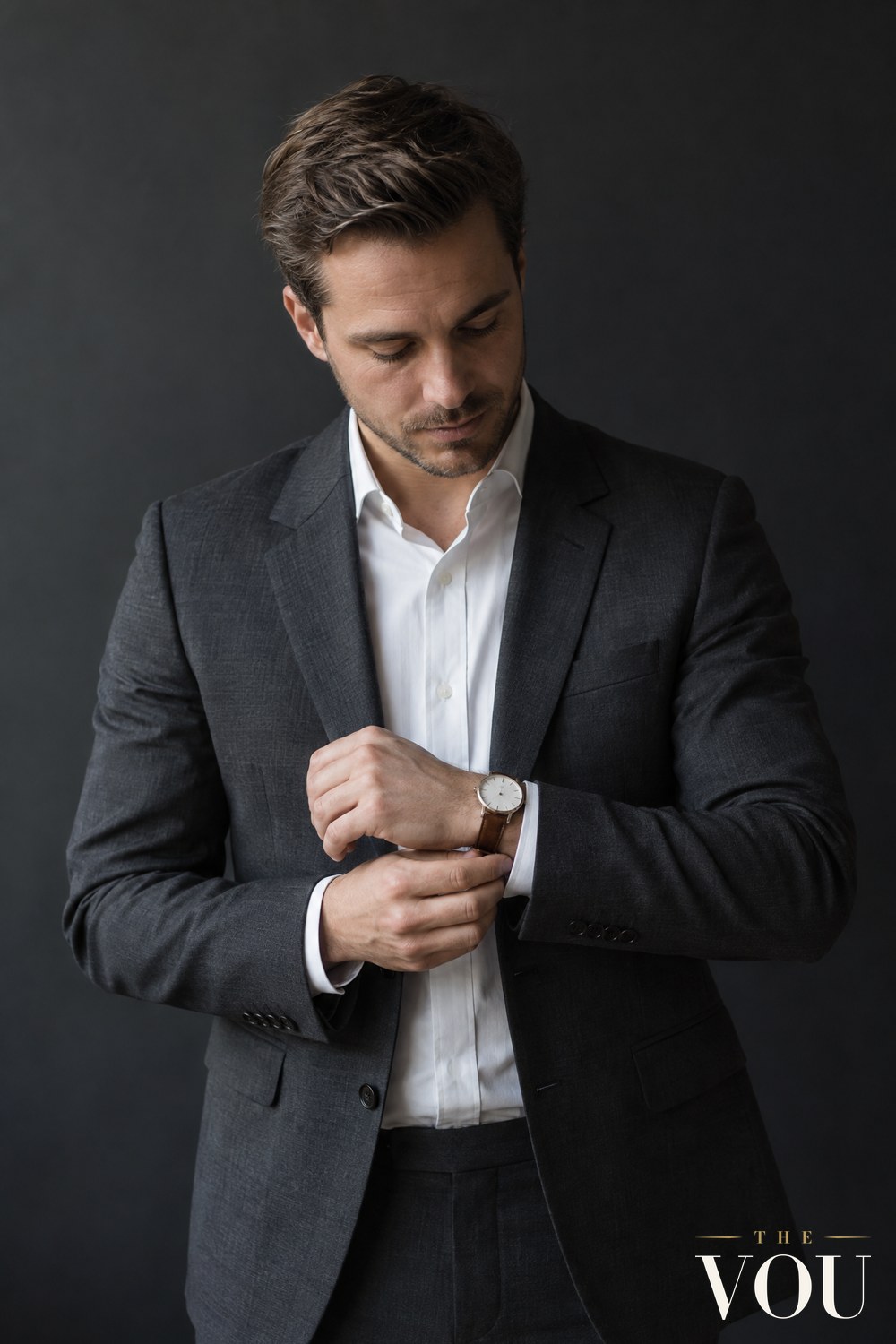A man in a charcoal suit jacket and white shirt adjusting a slim dress watch with a white dial and brown leather strap on his left wrist.