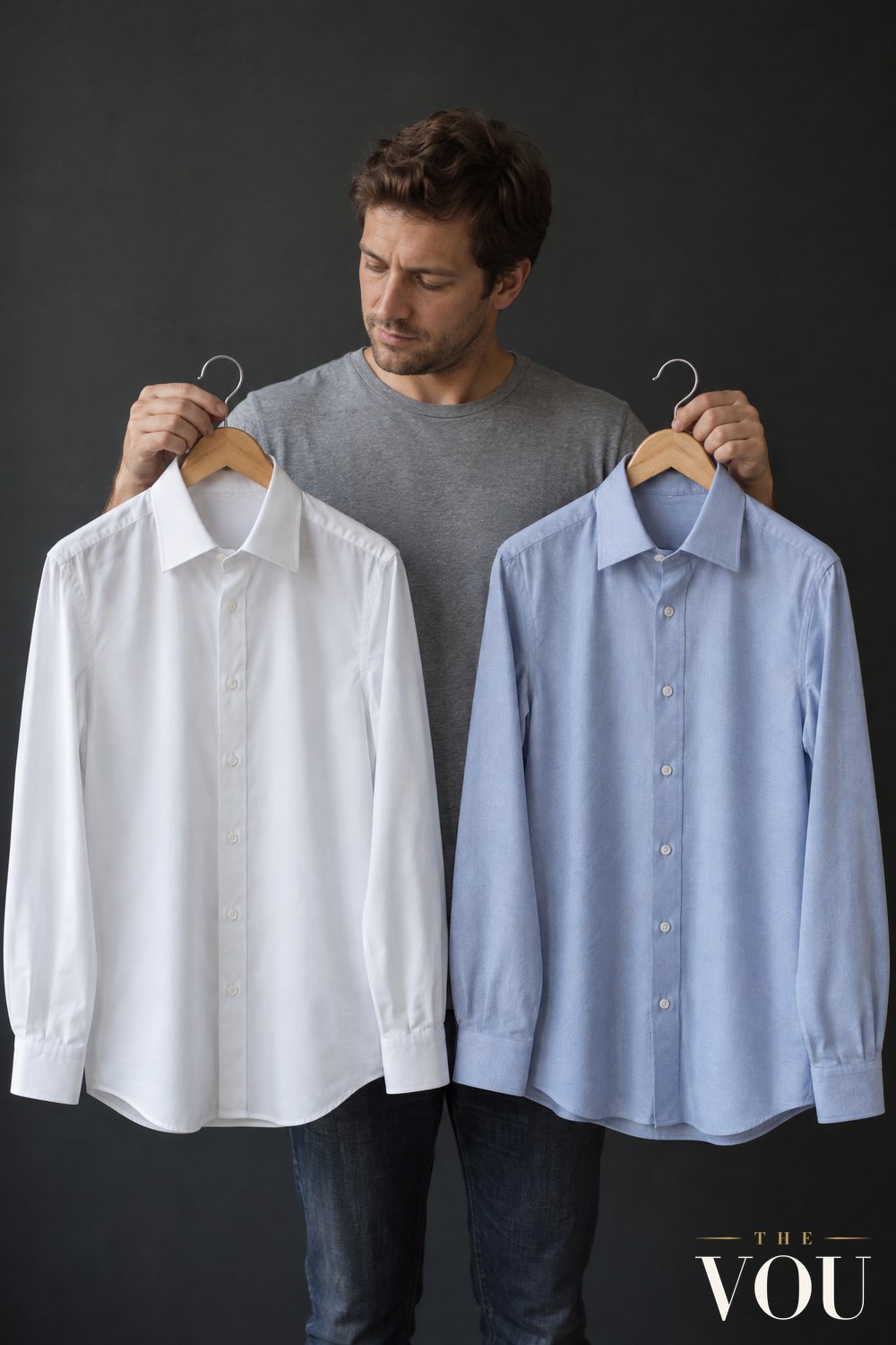 A man in a grey T-shirt holding up a smooth white poplin shirt and a light blue twill shirt on wooden hangers to compare the fabrics.