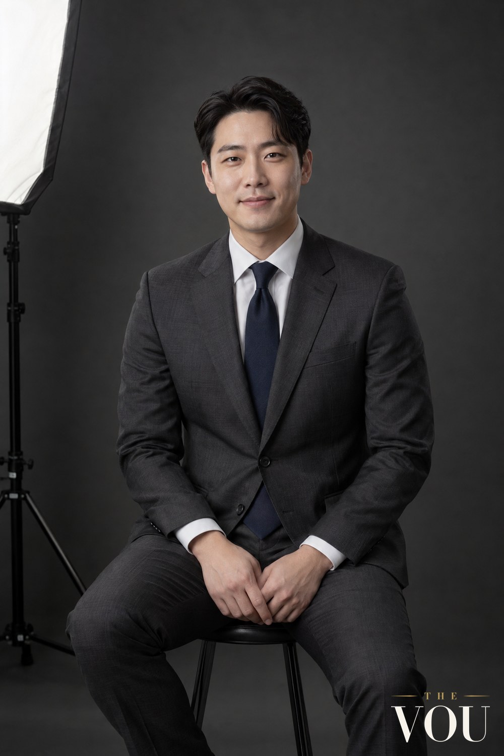 A man in a charcoal suit, white shirt and navy tie seated on a stool in a photographer's studio for his professional headshot.