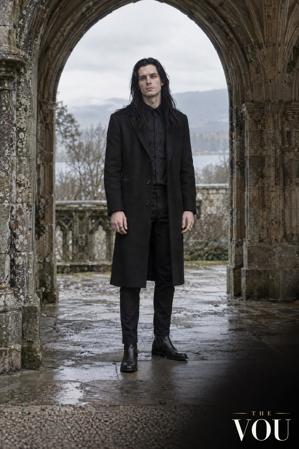 A man with long black hair in a long black wool coat, black shirt, black trousers and boots under a weathered gothic stone arch.