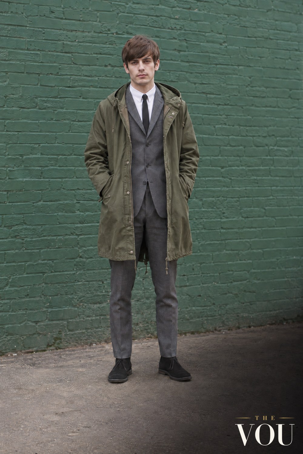 A young man with a mod haircut in a slim grey suit, white button-down shirt, thin black tie, olive fishtail parka and desert boots.
