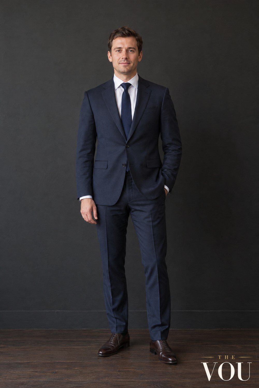 A man standing in a navy suit, white shirt and navy tie with dark brown leather Oxford shoes, against a charcoal wall.