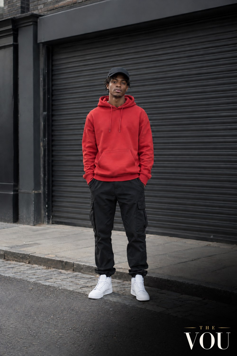 A young man in a plain red hoodie, black cap, black cargo trousers and white high-top trainers outside a shuttered streetwear shop.