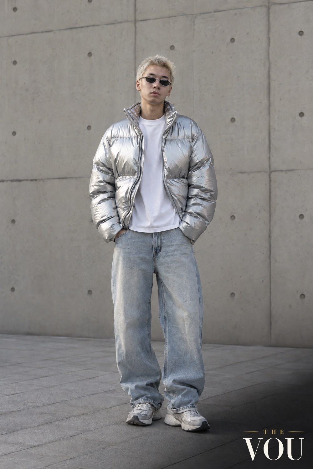 A young man with bleached hair in a silver metallic puffer jacket, white T-shirt, baggy light-wash jeans, small tinted sunglasses and chunky trainers.