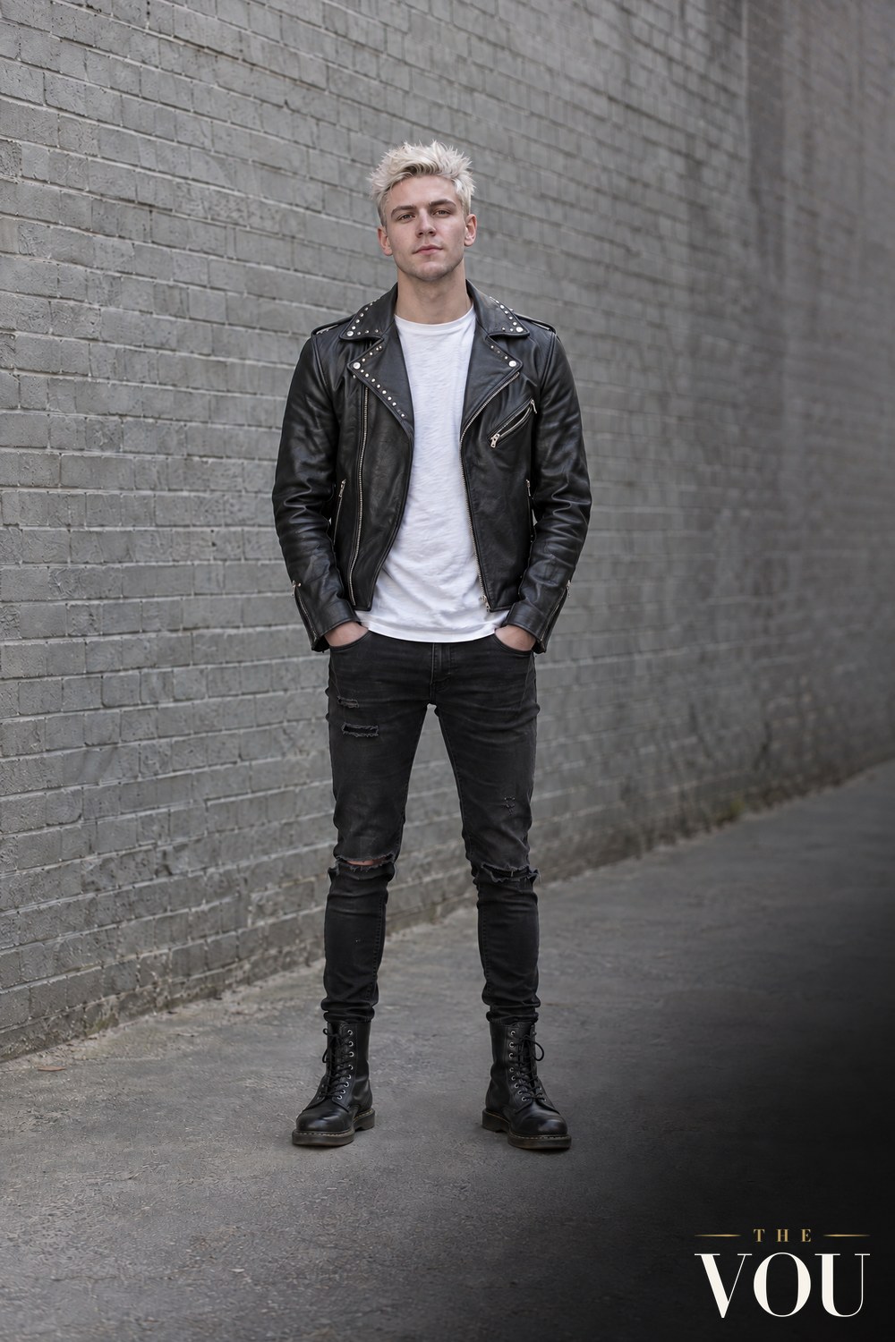 A man with spiky bleached hair in a studded black leather biker jacket, white T-shirt, ripped black skinny jeans and combat boots.