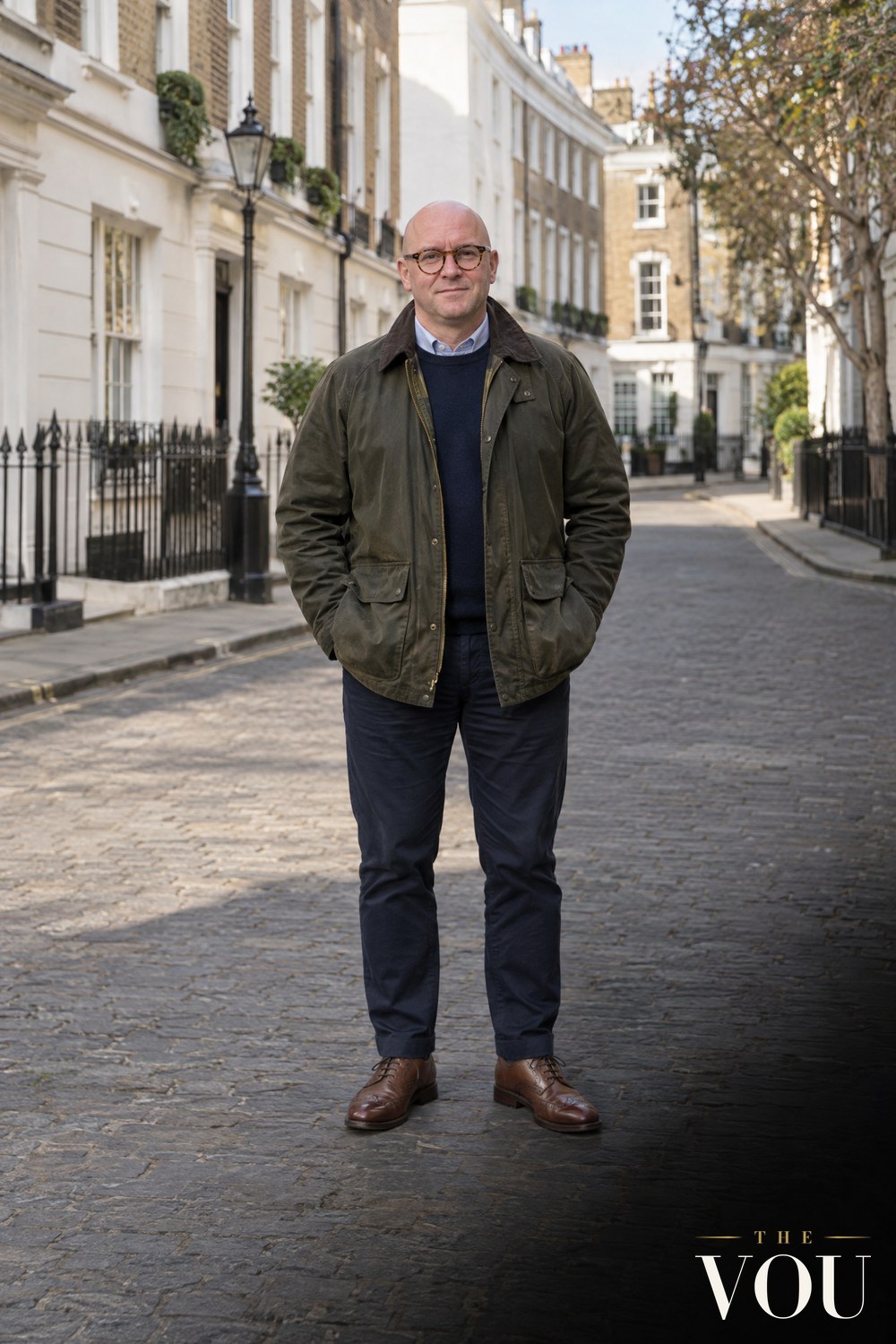 A bald man in round tortoiseshell glasses wearing an olive waxed cotton jacket, navy jumper, blue oxford shirt, navy chinos and brown leather shoes on a Georgian London street.
