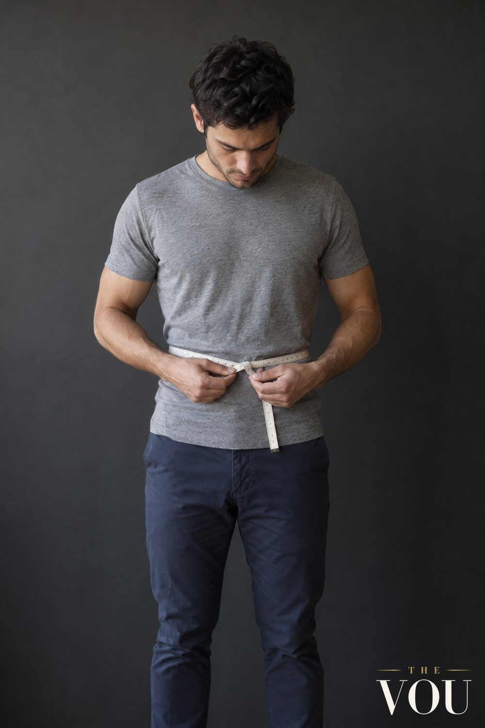 A man in a grey T-shirt and navy chinos holding a tailor's tape measure level around his natural waist.