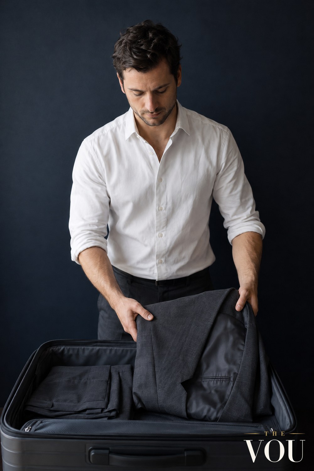 A man in a white shirt laying a charcoal suit jacket, folded in half with the lining out, into an open hard-shell suitcase beside folded trousers.