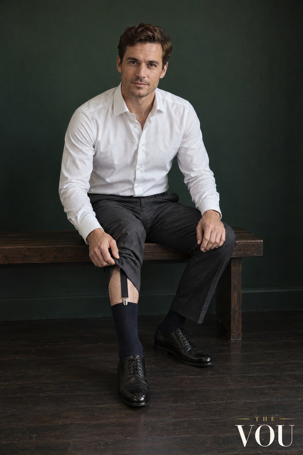 A seated man in a white shirt and charcoal trousers lifting one trouser leg to show a black elastic shirt stay clipped to the top of his navy sock.