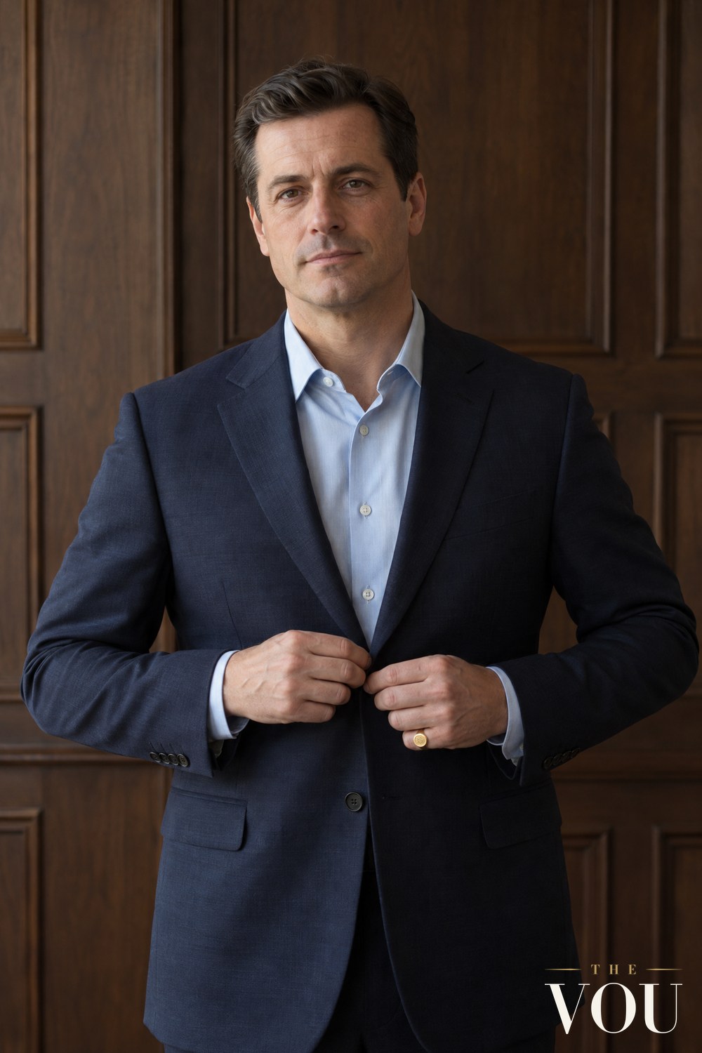 A man in a navy suit jacket and light blue shirt buttoning his jacket, a gold signet ring on the little finger of his left hand.