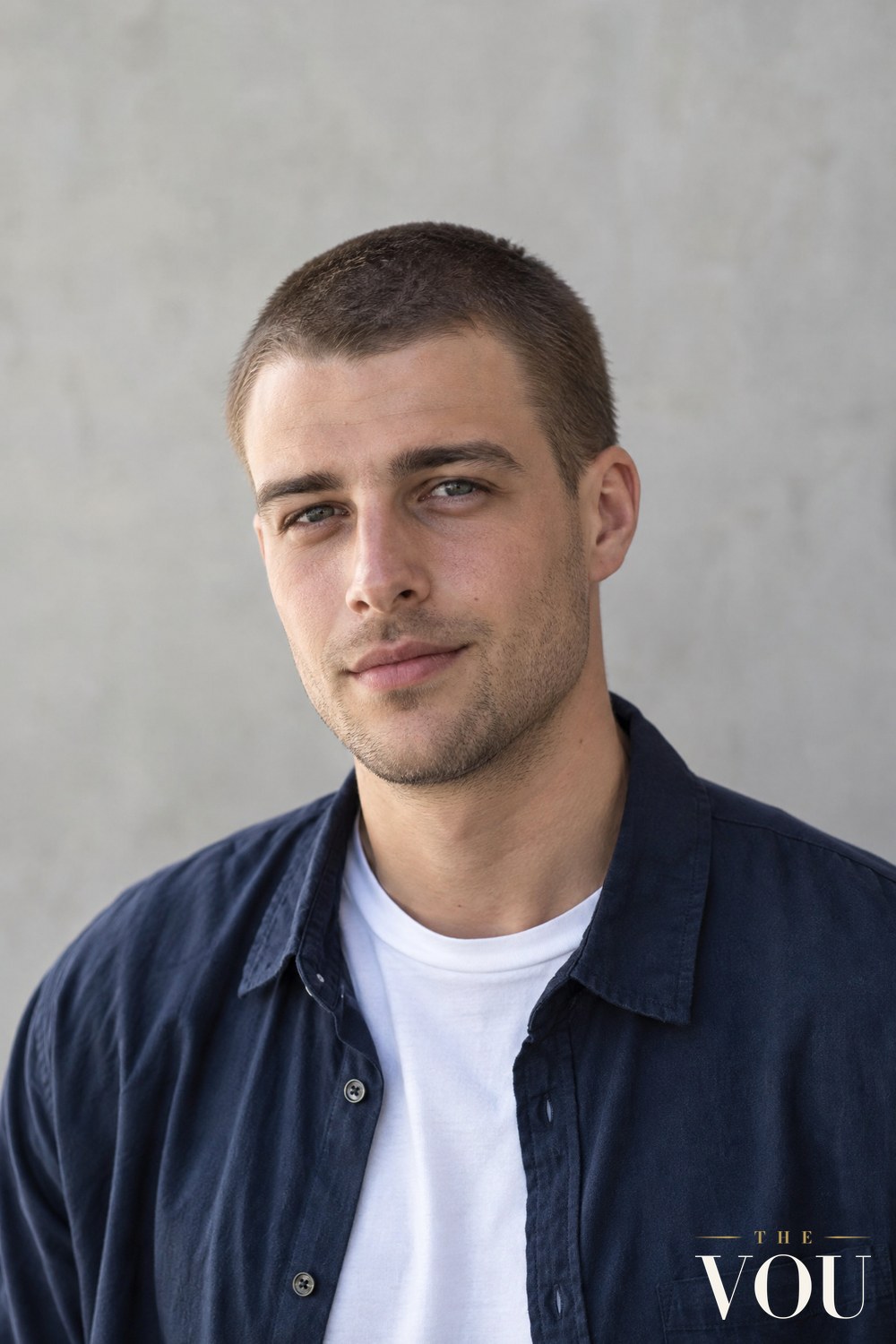 A man with an even buzz cut all over, wearing a white T-shirt and a navy overshirt against a pale grey wall.