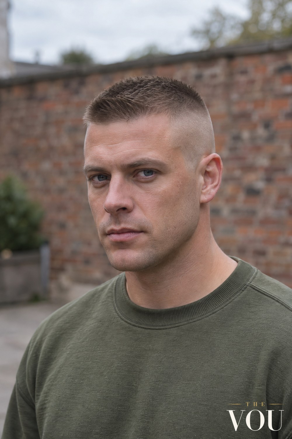 A man with a high and tight haircut, the sides and back clipped almost to the skin and a short flat patch on top, wearing an olive sweatshirt.