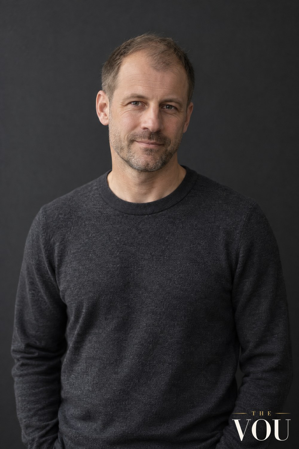 A man with thinning light brown hair cut in a short textured crop with low-contrast sides, in a charcoal jumper.