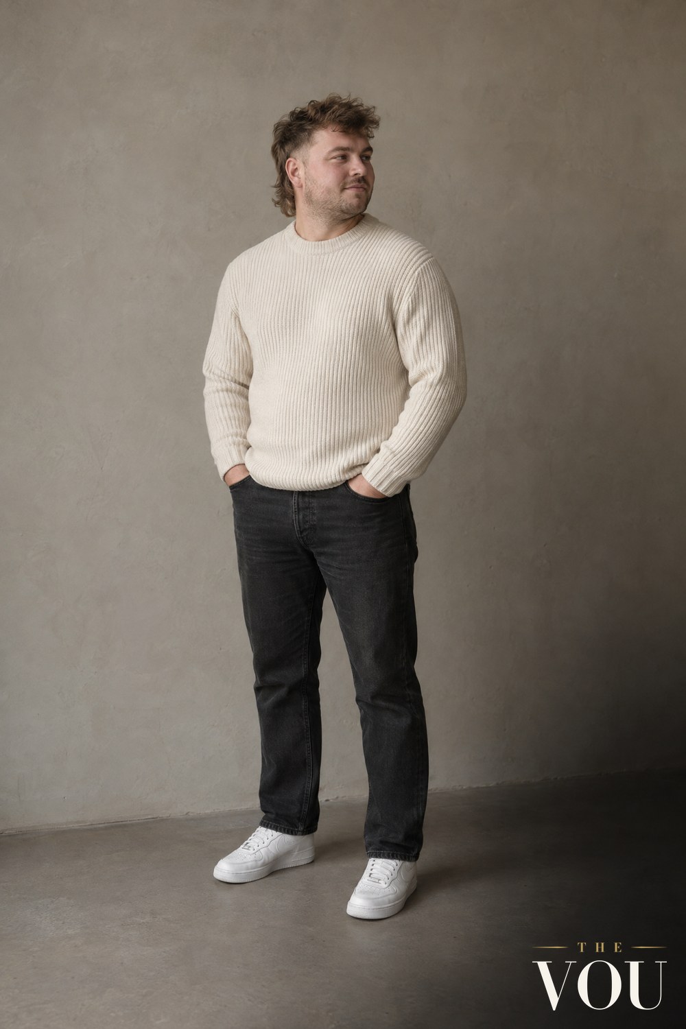 A man with a round face and a textured mullet, short at the sides and longer at the back, in a cream knitted jumper, dark jeans and white trainers.