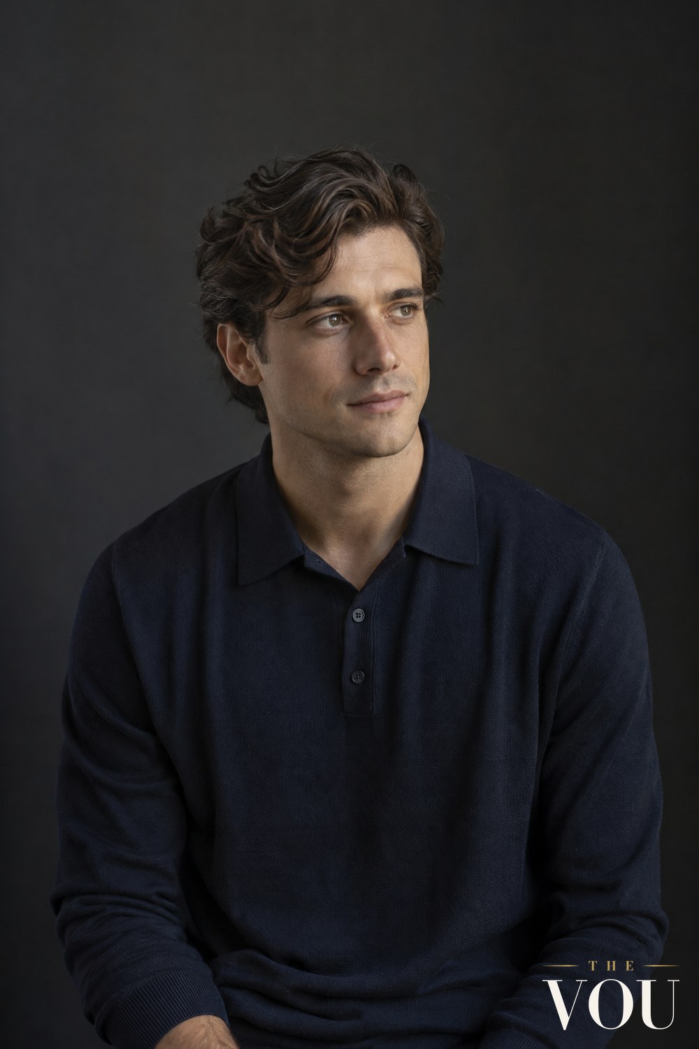 A man with medium-length dark brown wavy hair falling to one side, in a navy knit polo shirt, against a dark wall.