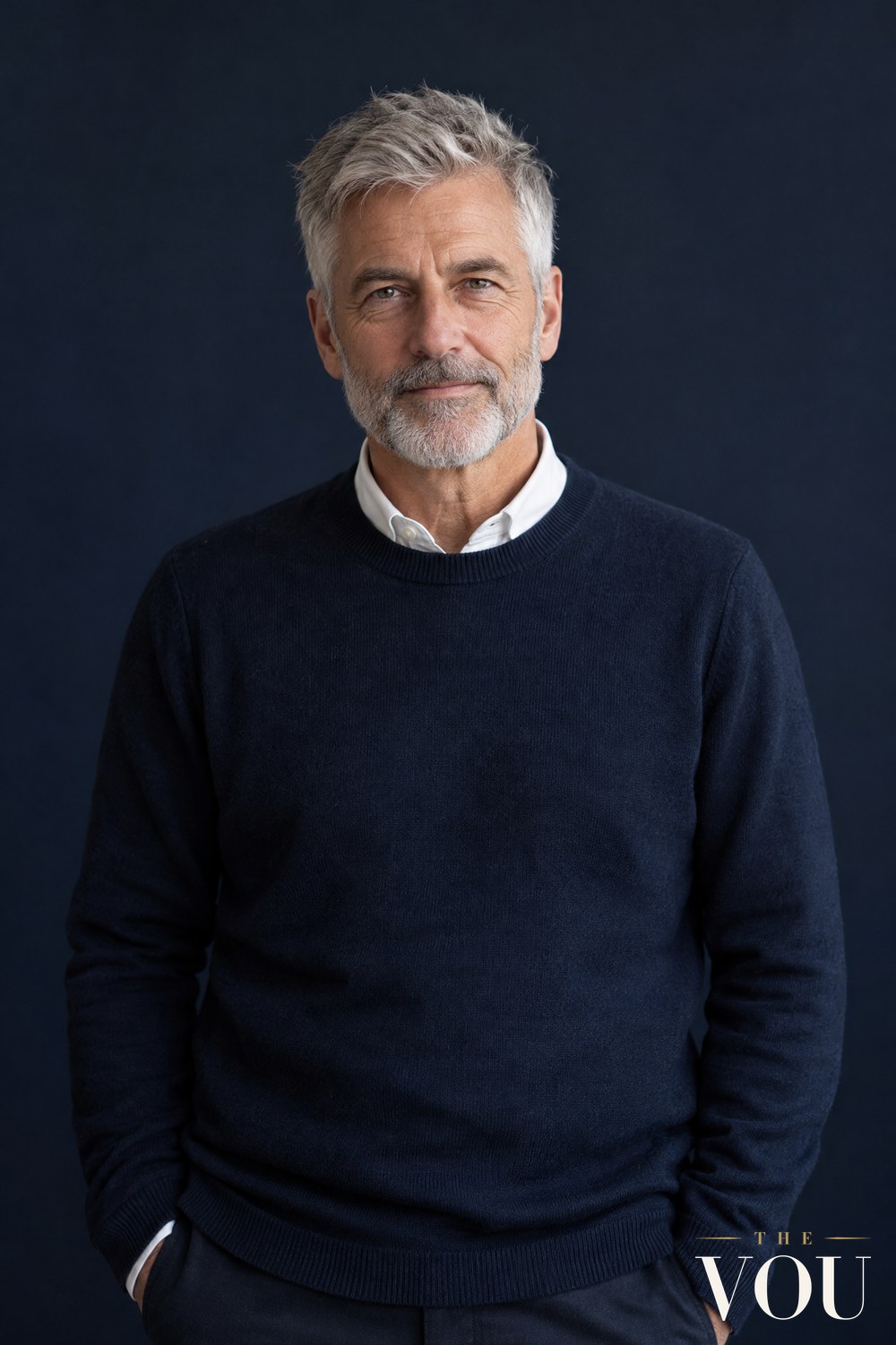A man in his fifties with a short textured crop of silver-grey hair and a trimmed grey beard, in a navy jumper over a white collar.